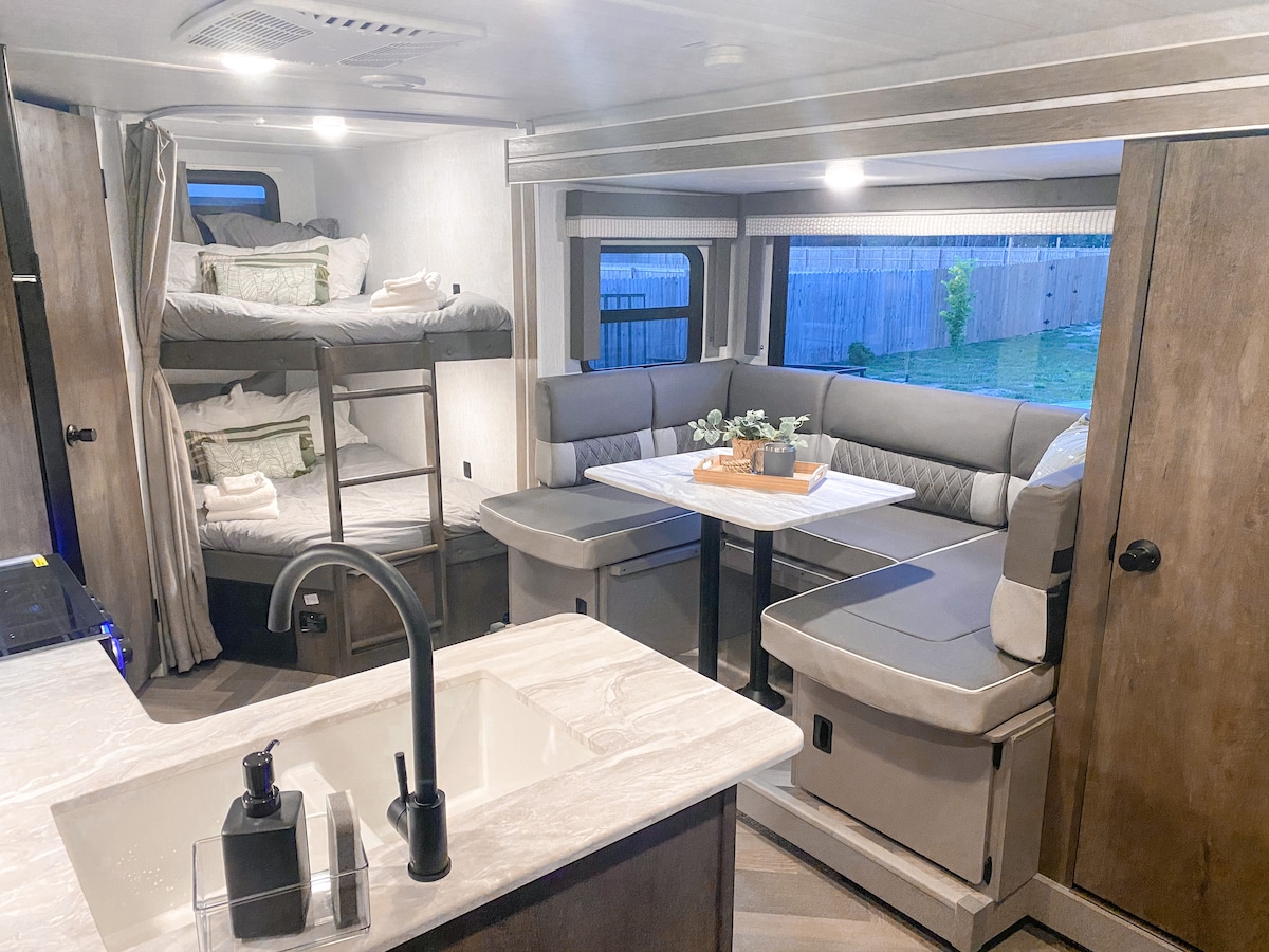 The interior of the camper reveals a cozy living area featuring a U-shaped dining booth with a table, a well-equipped kitchen area with a sleek sink, and two full-sized bunk beds. Natural light illuminates the space through a window, enhancing the inviting atmosphere.