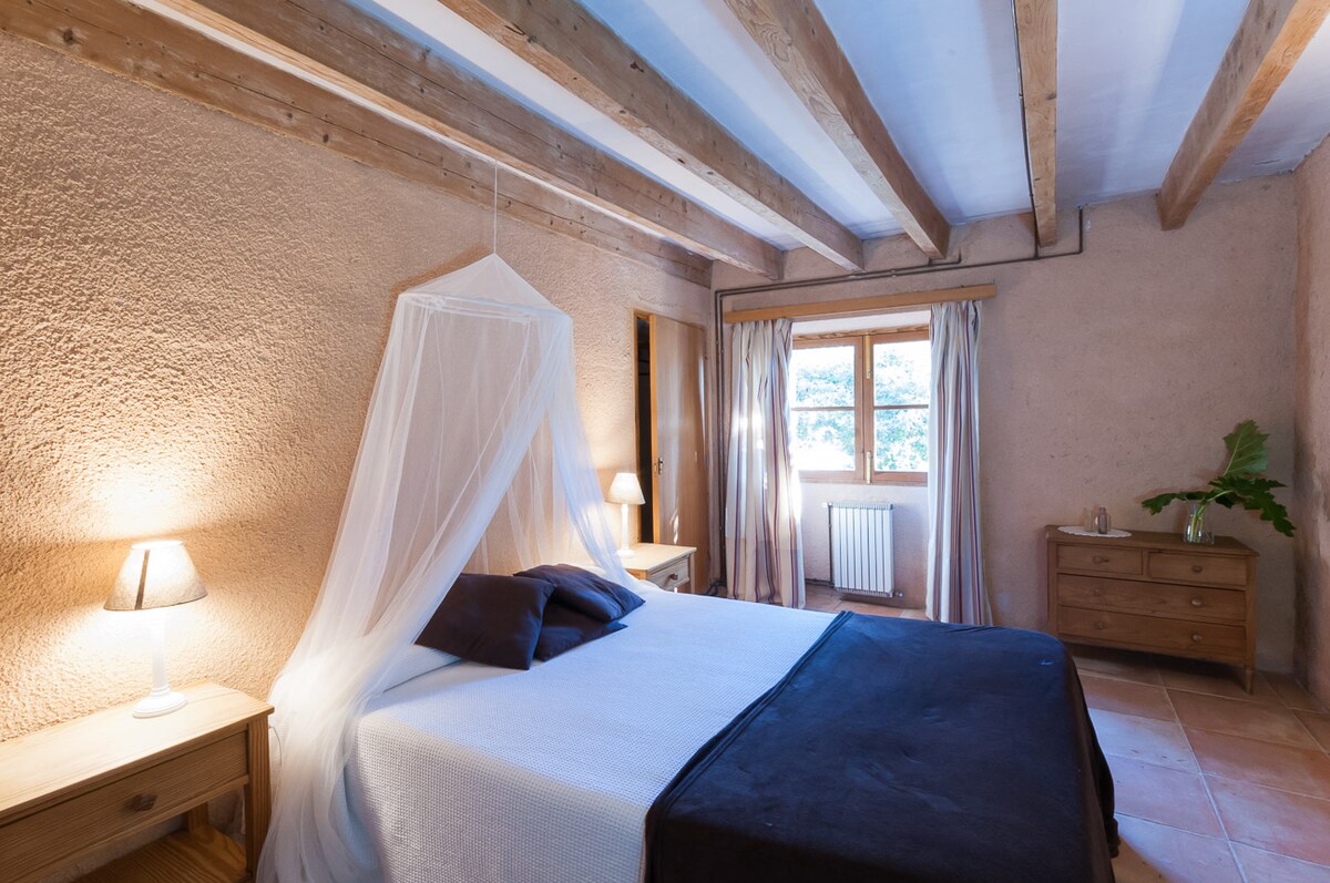 A spacious bedroom features exposed wooden beams and soft, earthy walls. A comfortable bed is dressed in crisp linens, accompanied by bedside tables with lamps. Natural light filters through a window, with light curtains offering a serene view. A wooden dresser provides additional storage.