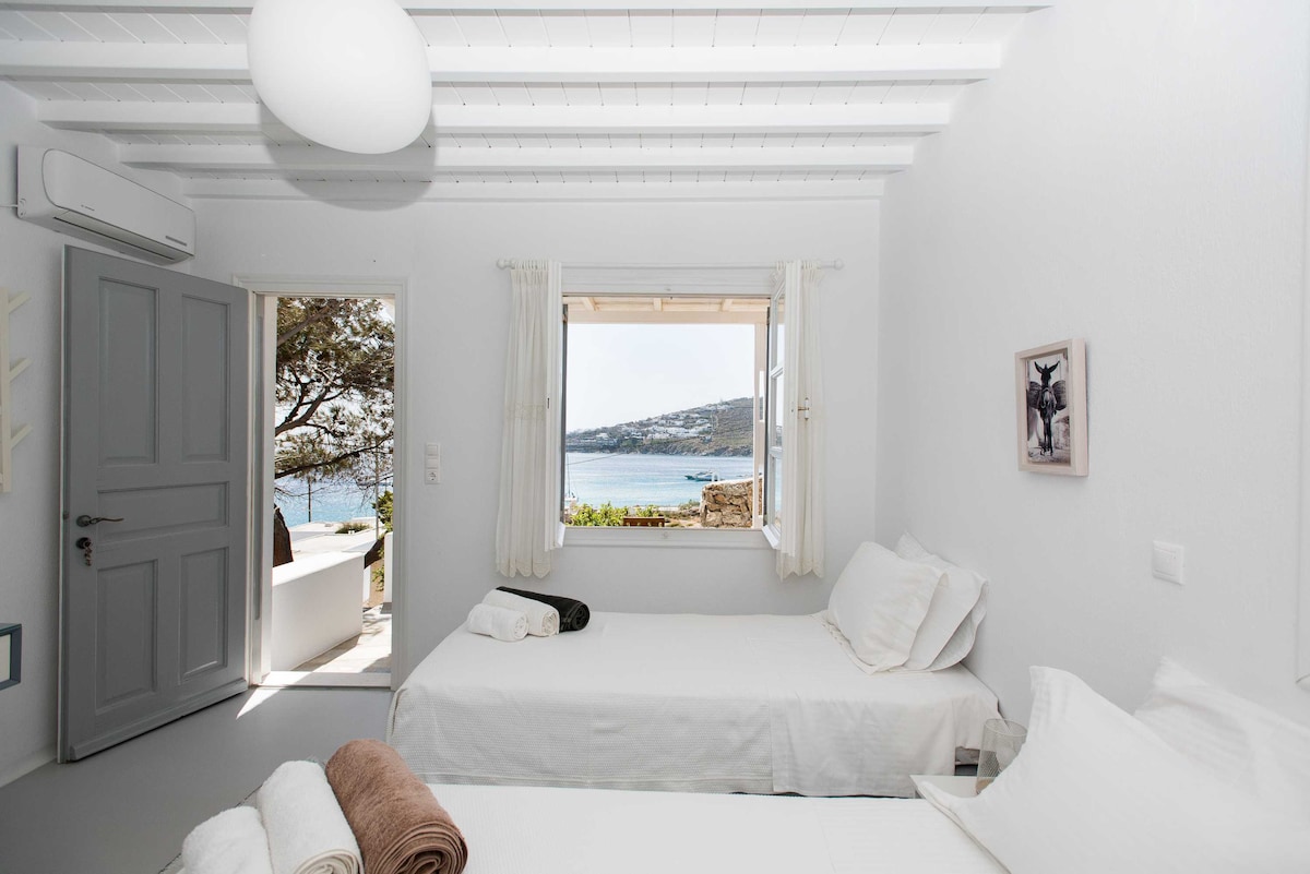 A bright guest room features two spacious beds, with views of the blue waters visible through large windows. The light-colored walls and ceiling reflect natural light, while a door leads to a private veranda. Soft furnishings include towels neatly arranged at the foot of each bed.