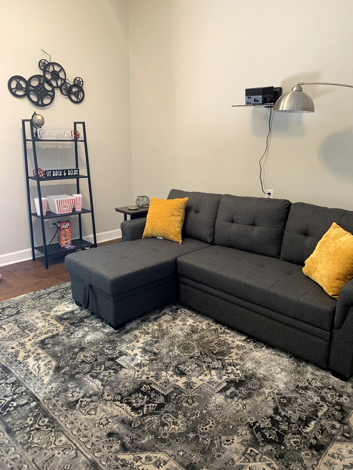 A comfortable gray sectional sofa with yellow accent pillows is positioned on a patterned area rug. A small bookshelf nearby displays popcorn and movie-themed items, while a modern lamp provides soft light. A media player is mounted on the wall, enhancing the cozy atmosphere.