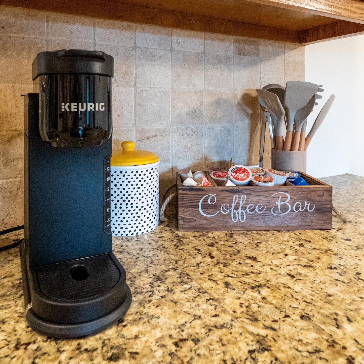 A coffee station is presented with a Keurig machine positioned on a granite countertop. A wooden box labeled 'Coffee Bar' holds various coffee pods and supplies. Kitchen utensils are neatly organized in ceramic and wooden containers, complementing the overall setup.