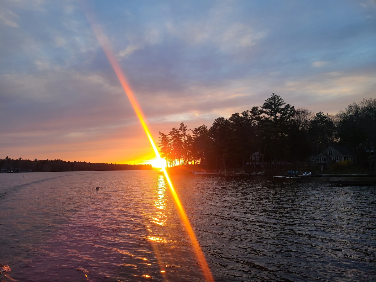 A vibrant sunset casts golden rays over the water, reflecting off the expansive lake. Silhouetted trees line the shore, while distant homes are faintly visible. The sky transitions from warm orange to soft purple hues, creating a serene ambiance over the landscape.