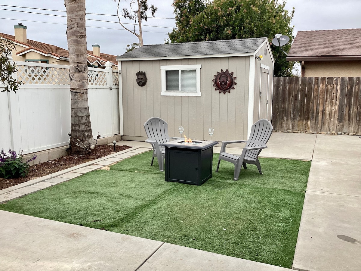 A private outdoor space features a small green lawn area with two light gray Adirondack chairs arranged around a fire pit table. A shed is visible in the background, with a window and decorative wall ornament. A white fence and palm trees complete the inviting setting.
