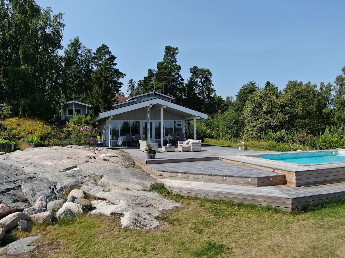 A wooden villa is set among towering trees, featuring a private pool surrounded by a wooden deck. Natural stone elements create a seamless transition between the landscaped area and the pool. Sunlight casts a warm glow on the exterior, highlighting its inviting architecture.