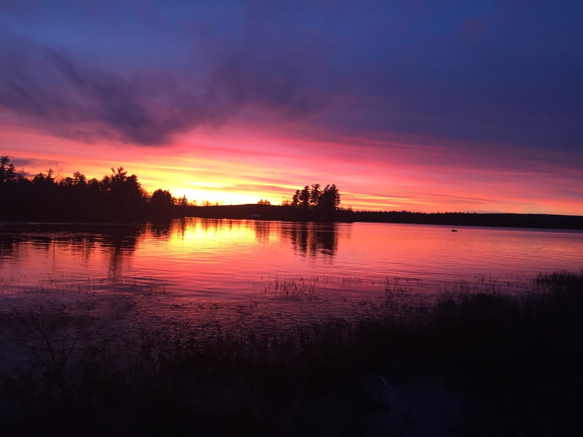 A vibrant sunset casts hues of orange, pink, and purple across the lake, reflecting softly on the water's surface. Silhouettes of trees line the horizon, enhancing the serene atmosphere of the rural setting.