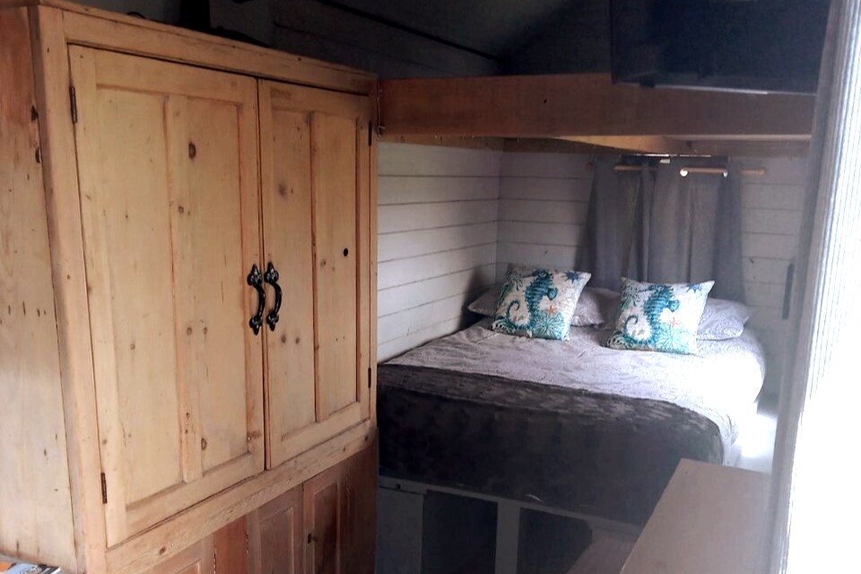 An interior view showcases a cozy sleeping area featuring a spacious bed dressed in neutral linens. Two decorative pillows with a blue design are positioned on the bed. A wooden cupboard with double doors stands beside the bed, contributing to the rustic charm of the space.