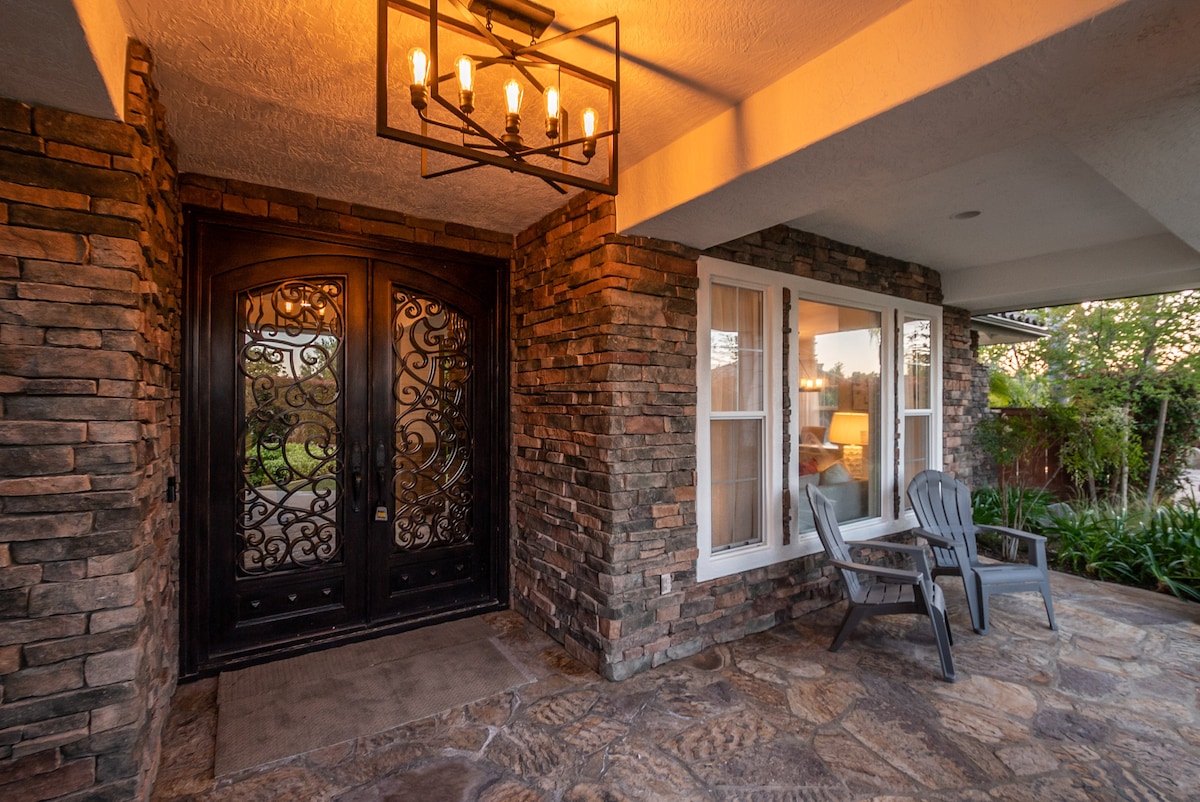 A welcoming entrance features two large, ornate doors of dark wood adorned with intricate ironwork. Adjacent, two distinctive chairs are positioned on a stone patio. Soft lighting from a chandelier illuminates the area, while lush greenery provides a natural backdrop.