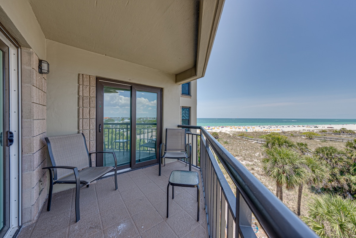 A balcony is shown, featuring two metal chairs and a small table, with views of the beach and ocean visible in the background. Palm trees and sandy areas can be seen through the railing, enhancing the coastal atmosphere.