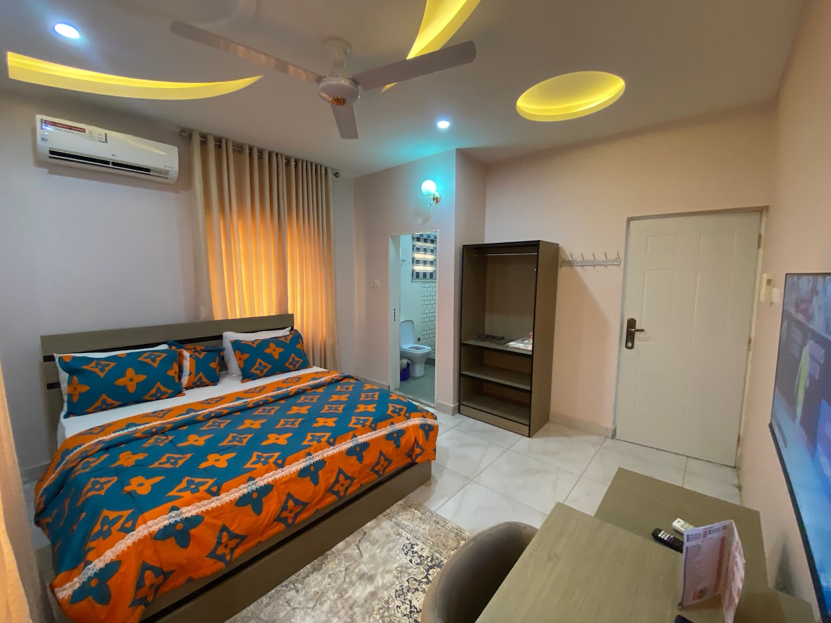 The suite features a spacious bed adorned with vibrant orange and blue bedding, complemented by light-colored walls. A ceiling fan is present, and a television is mounted on the wall. A kitchenette area is visible, along with a seating arrangement and bathroom access.