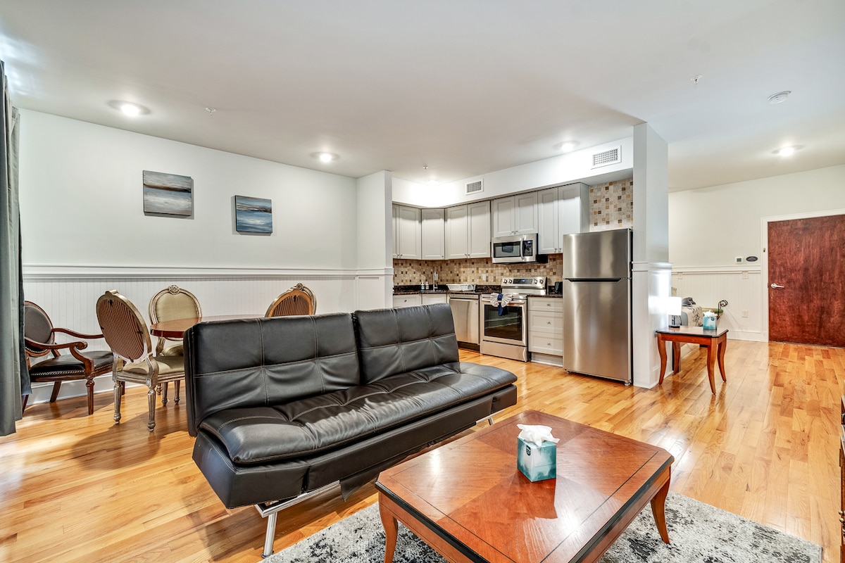 A modern studio apartment features a comfortable black sleeper sofa, a wooden coffee table, and a spacious dining area with elegant chairs. The kitchen is equipped with stainless steel appliances and light cabinetry. Natural light brightens the polished hardwood floors, enhancing the welcoming atmosphere.