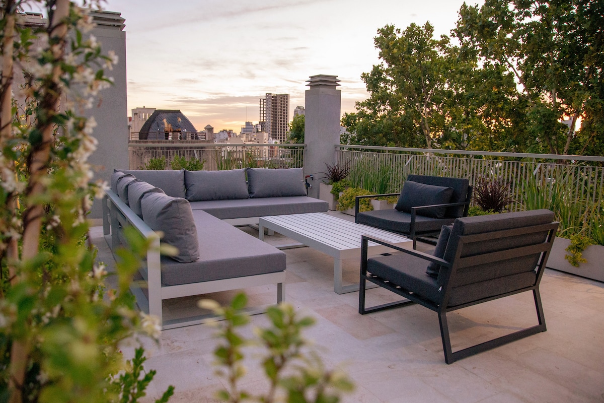 A rooftop terrace features a spacious seating area with gray cushions surrounding a low coffee table. Lush greenery lines the perimeter, providing a serene atmosphere against the backdrop of the city skyline at sunset.