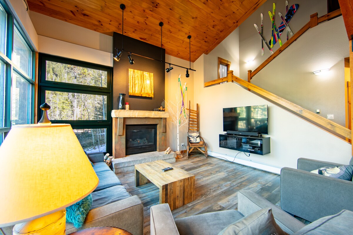 The living area features an extensive wall of windows, providing views of the outdoors. A comfortable seating arrangement includes a plush sofa and chairs. A natural wood coffee table is centered near a gas fireplace, and a large flat screen TV is mounted on the wall. Ski decor adds a playful touch.