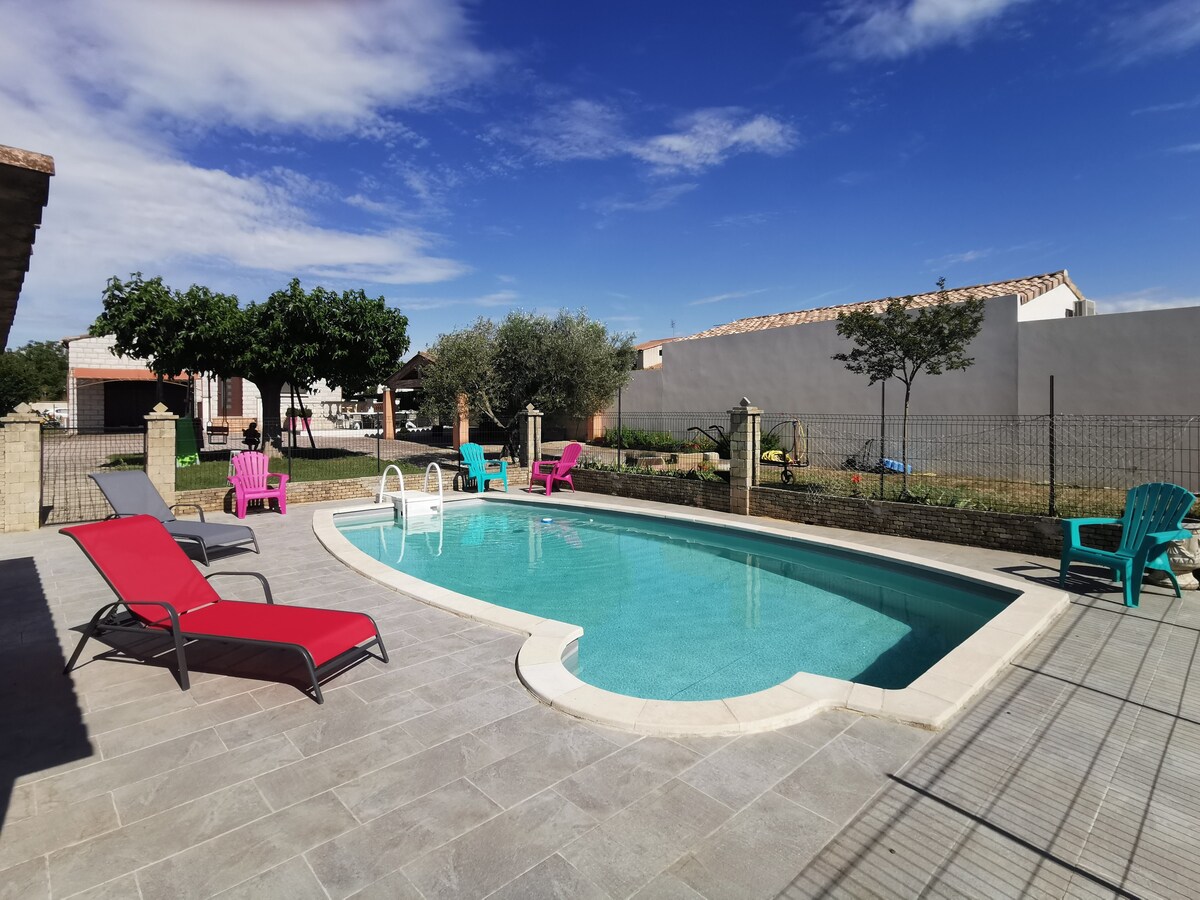 A private swimming pool features clear water and is surrounded by a variety of colorful lounge chairs. Landscaping includes trees and a manicured lawn, with a fence providing privacy. The setting is illuminated by natural light under a blue sky.