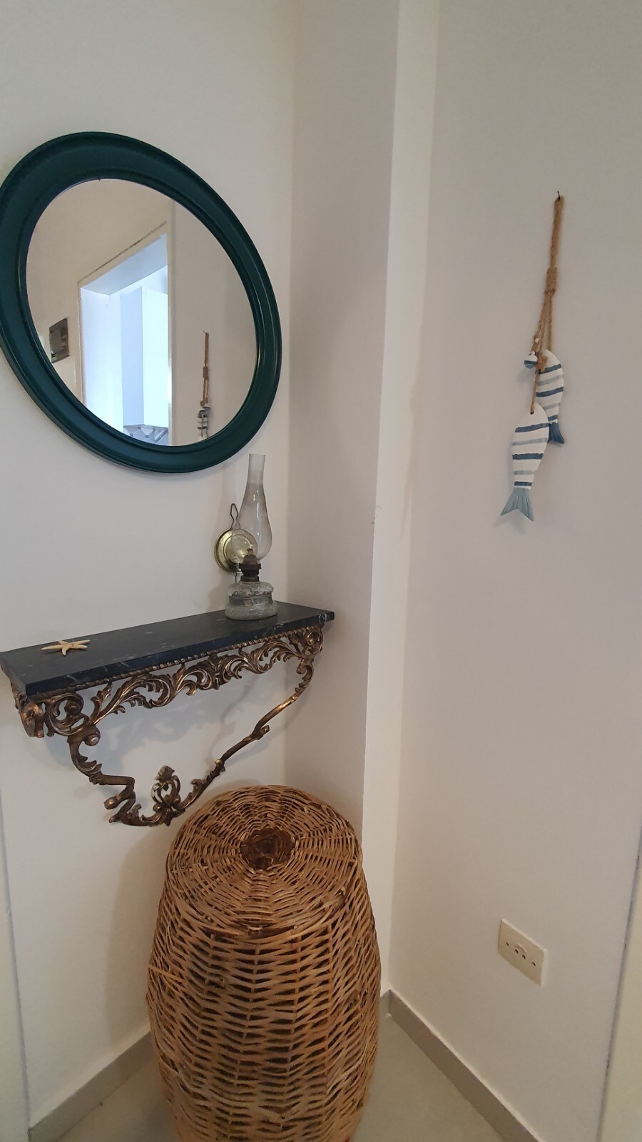A corner of the entrance features a decorative wall-mounted shelf with an antique lamp and a round mirror framed in teal. A woven basket sits below, adding a natural texture to the space. Simple nautical decor hangs nearby, enhancing the coastal theme.