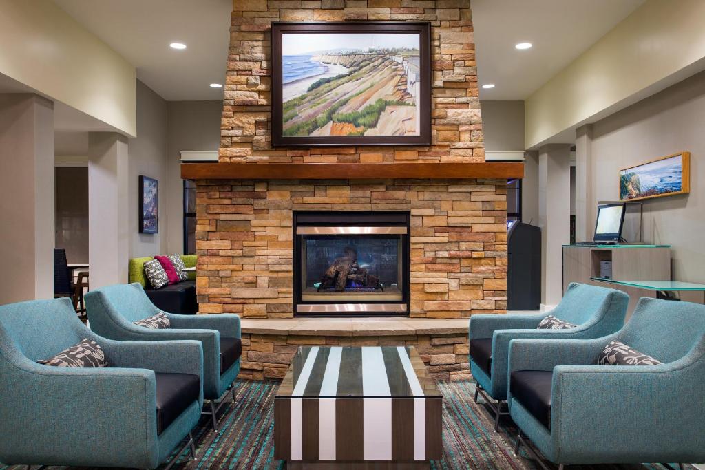 A warm and inviting seating area showcases four upholstered chairs arranged around a striped coffee table. A stone fireplace is centered along the wall, topped with a landscape painting that depicts coastal scenery. Soft lighting enhances the comfortable ambiance.