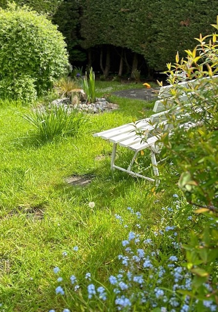 A serene garden setting is shown, featuring a white metal bench positioned on a lush green lawn. Colorful flower beds are visible, complemented by tall shrubs and a small water feature, creating a tranquil outdoor space.