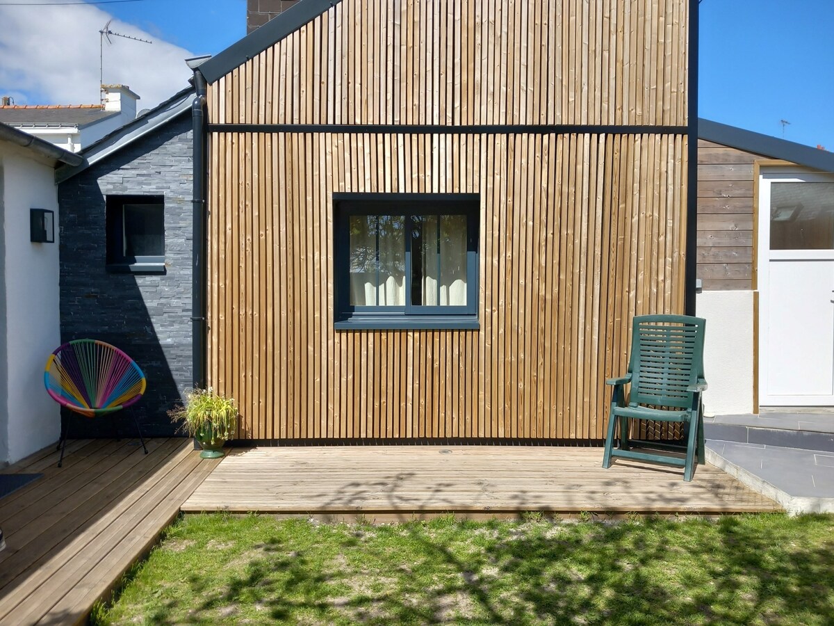 The exterior of the house features a combination of wood and stone materials, with vertical wooden slats accentuating a central window. A green chair sits on a wooden deck in front of a small grassy area, complemented by a potted plant.