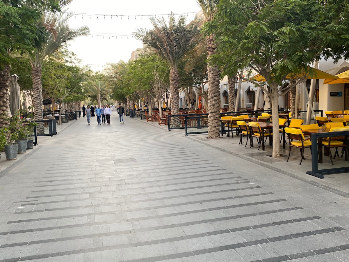 A wide promenade lined with palm trees features a path marked by decorative stripes. Outdoor seating is visible along the sides, with yellow chairs and tables arranged under shaded canopies. Groups of people are seen strolling, creating a vibrant atmosphere.