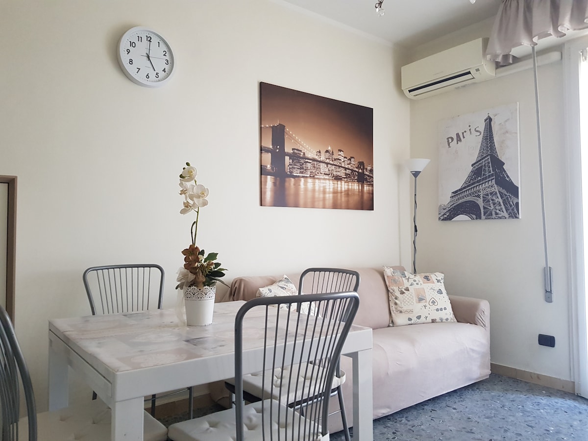 A cozy living area is displayed, featuring a light-colored sofa and a dining table with four chairs. A wall clock and framed artwork depicting a cityscape add character, while a potted plant sits as a centerpiece. Natural light streams in from a nearby window.