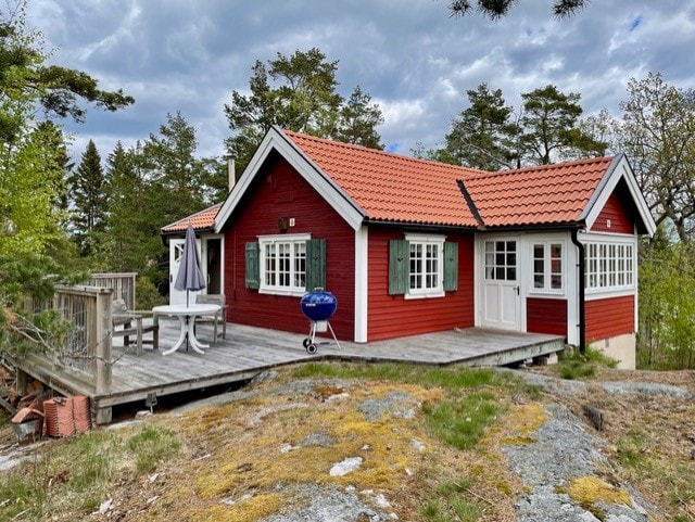 A charming red cottage is nestled among greenery, featuring a sloping roof and white trim. A spacious wooden deck extends from the cottage, with outdoor seating and a small grill visible. Surrounding trees frame the house, creating a peaceful retreat in the landscape.