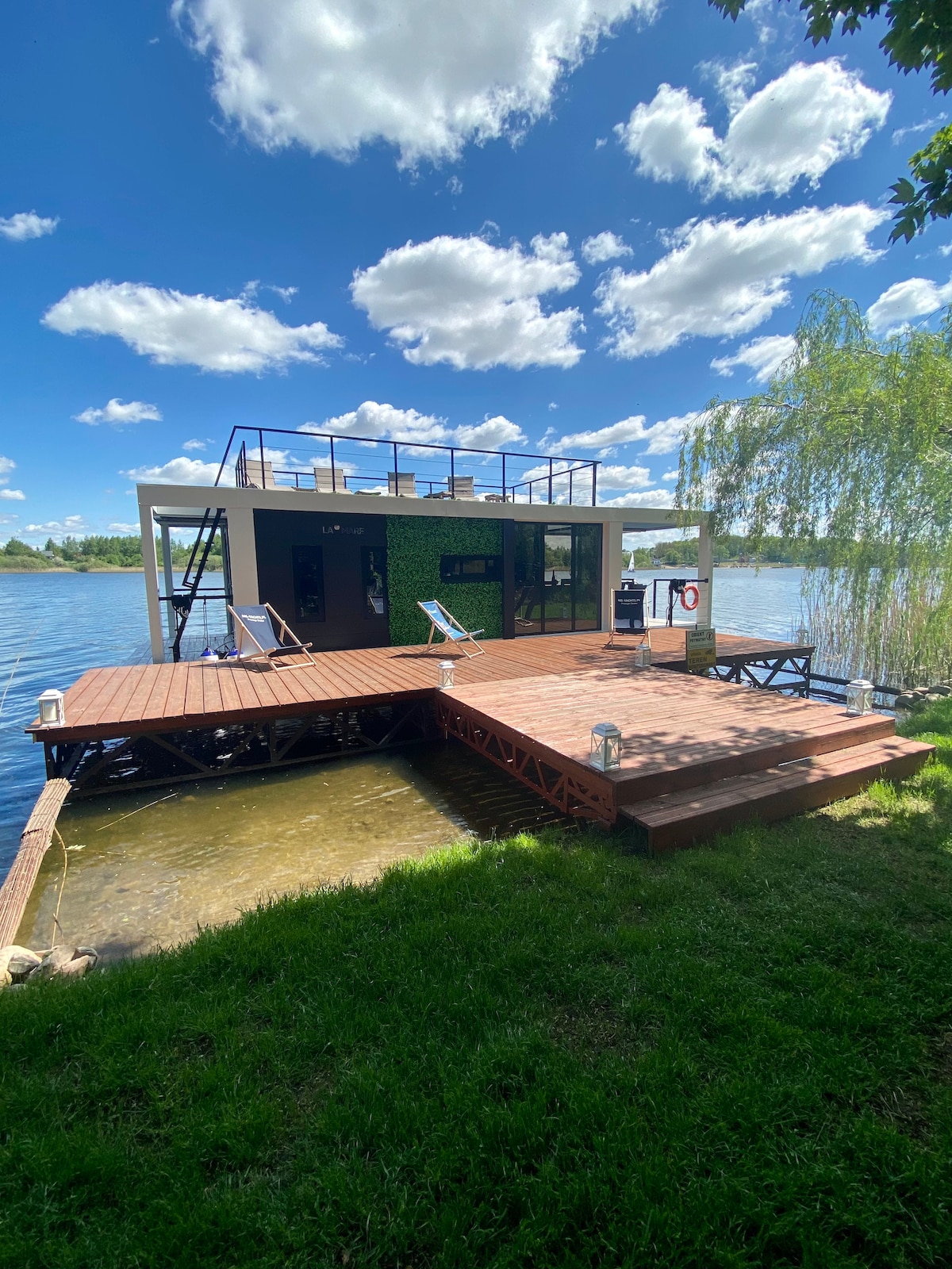 A modern houseboat is positioned on a lake, featuring a wooden deck that leads to the water. Lush green grass surrounds the structure, while fluffy white clouds drift in the vibrant blue sky. A spacious upper terrace is visible, offering a scenic view.