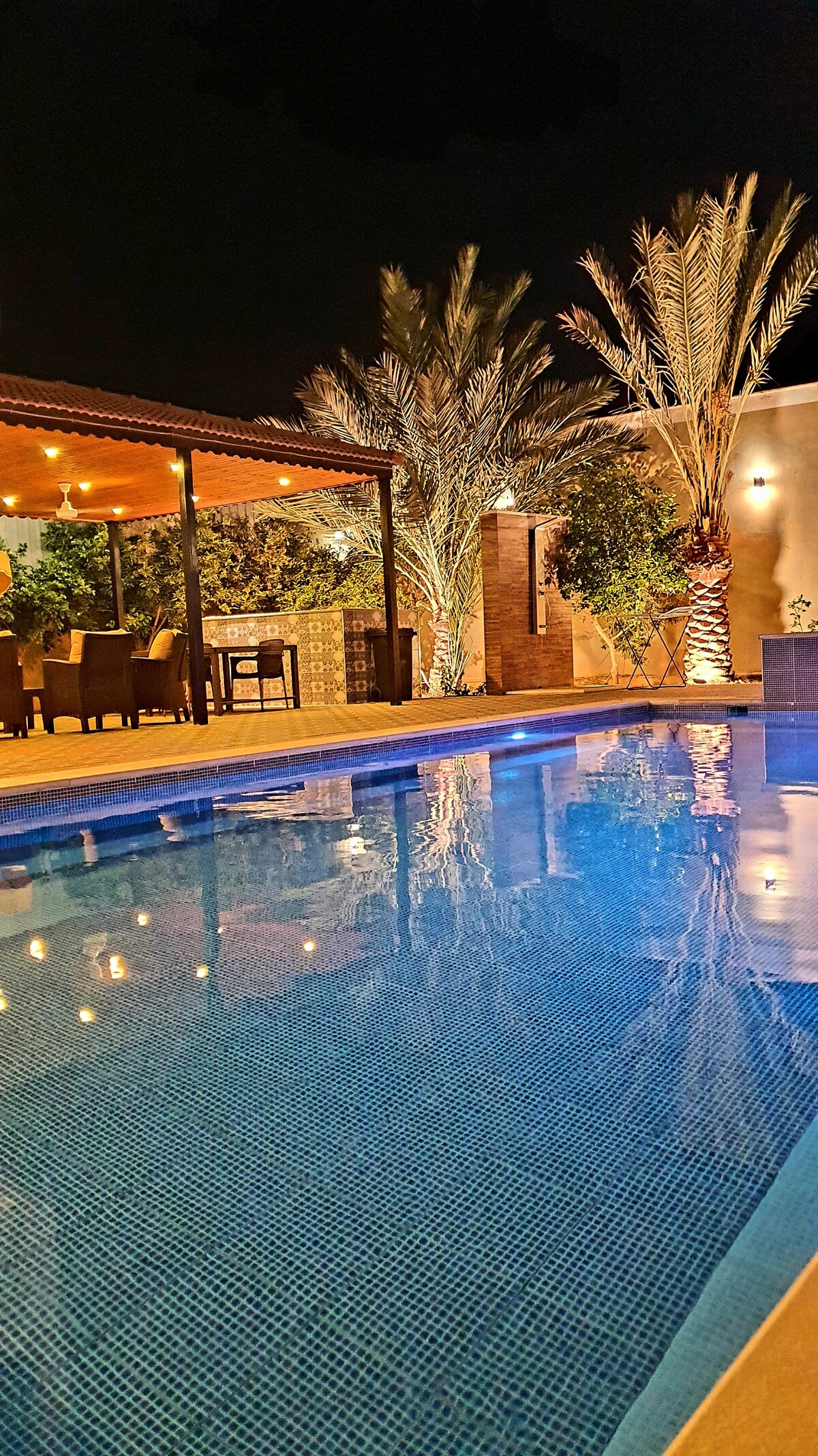 A tranquil swimming pool glows under soft lighting, reflecting the surrounding palm trees and outdoor dining area. A shaded seating area is positioned nearby, creating an inviting space for gatherings. The warm atmosphere is complemented by decorative landscaping and ambient lighting.