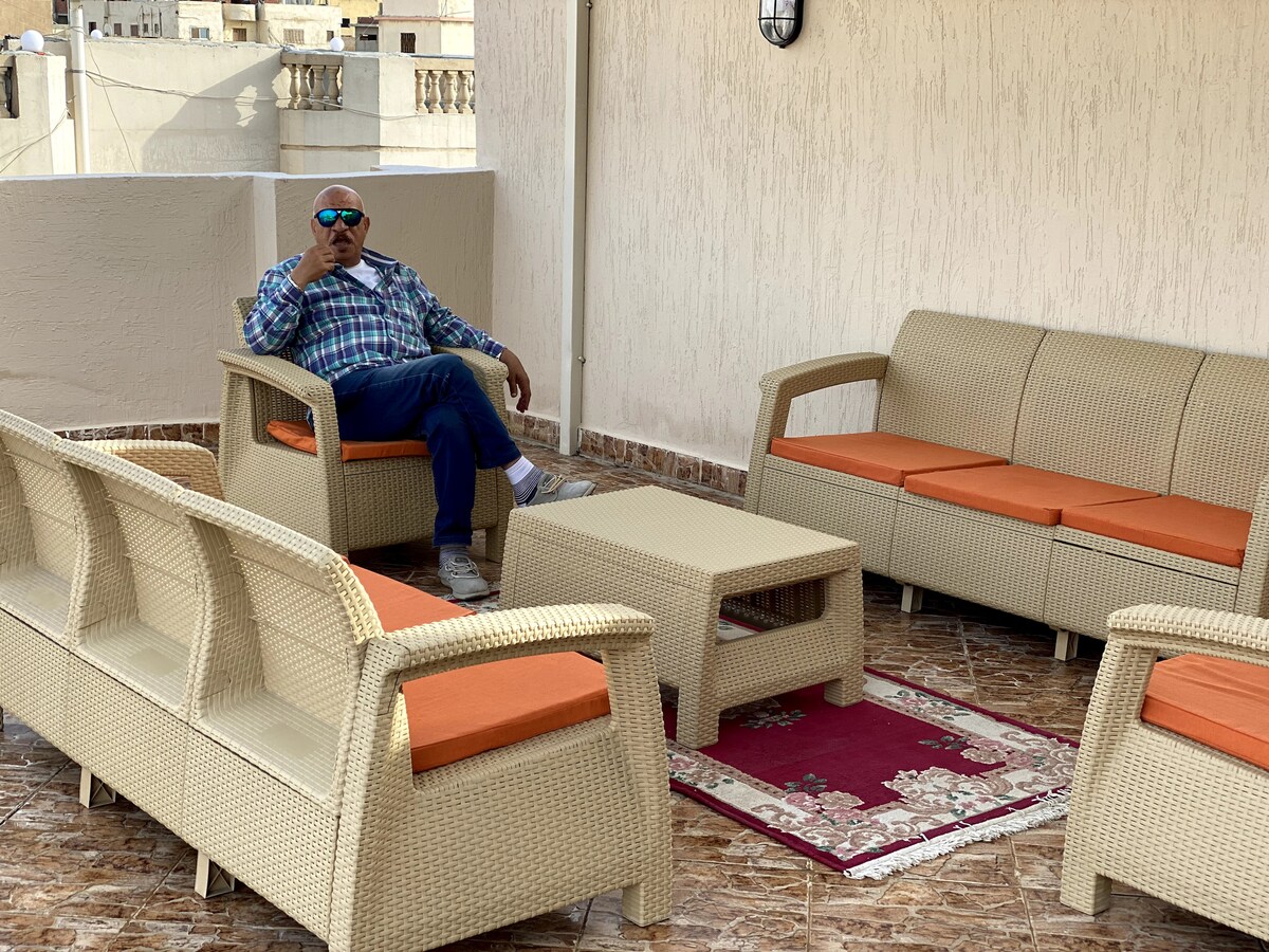 A furnished terrace features a seating area with light-colored rattan furniture, including two sofas and four armchairs, complemented by orange cushions. A low coffee table sits in the center, resting on a decorative rug. Sunlight bathes the space, creating a relaxed atmosphere.