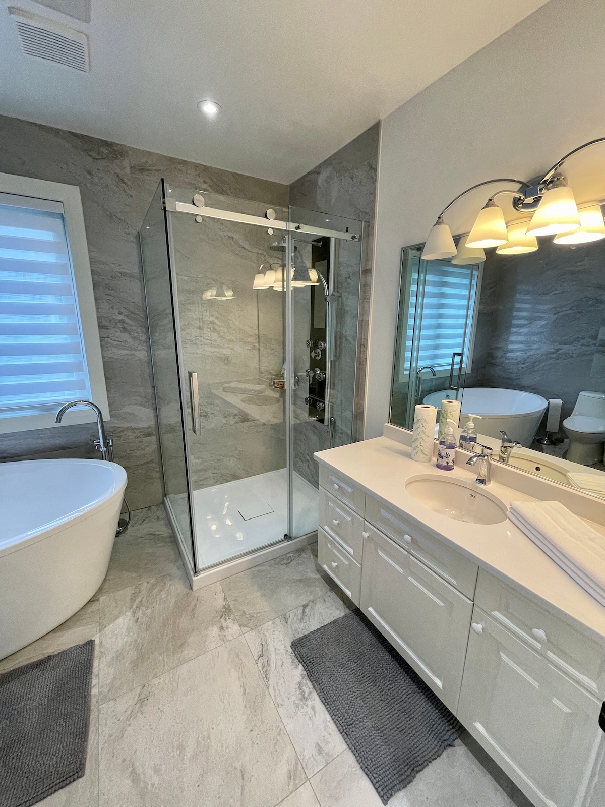 The bathroom features a sleek glass shower enclosure next to a modern freestanding tub. A double vanity with ample countertop space is complemented by a large mirror. Soft lighting is provided by a fixture above, enhancing the neutral tones of the tiled walls and flooring.