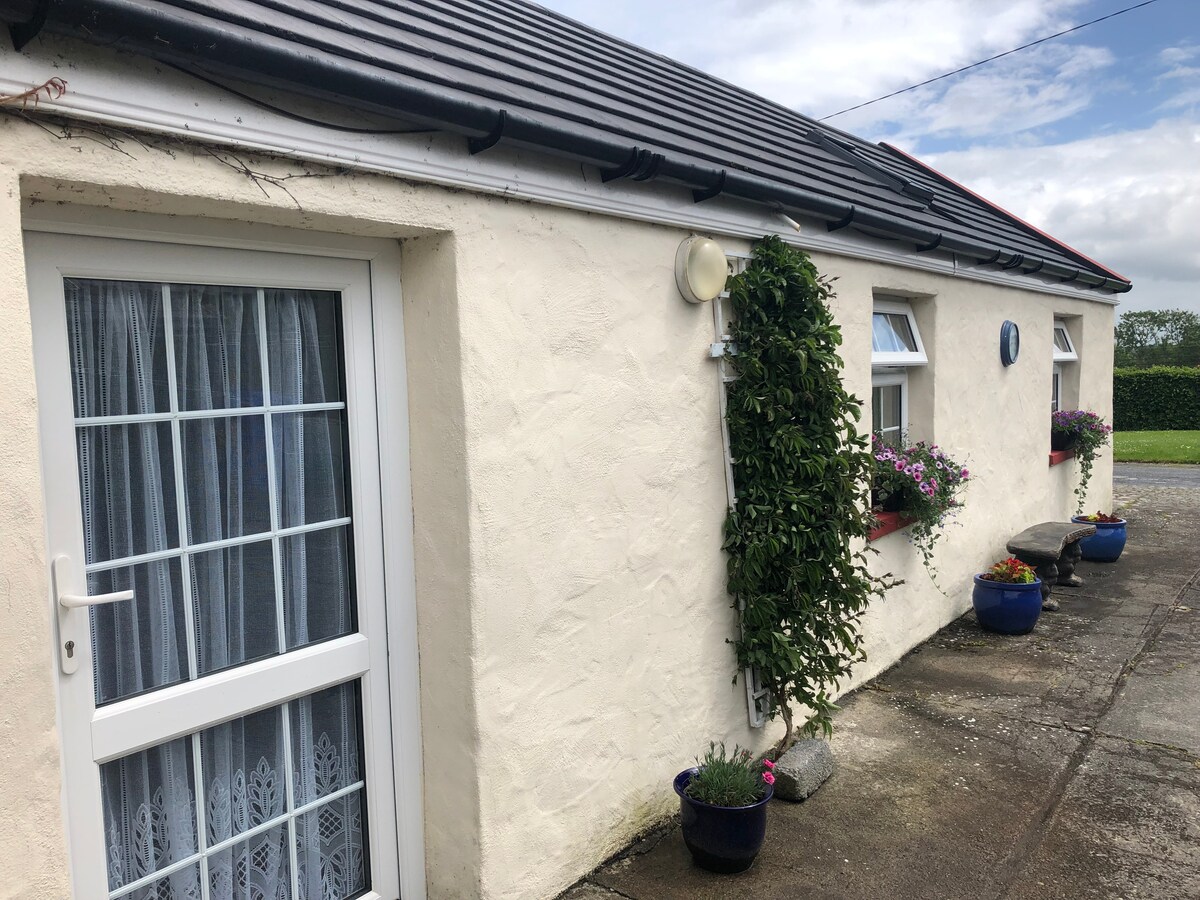 The exterior of a quaint cottage is displayed, featuring a light-colored façade with decorative plants adorning the walls. A welcoming entrance is framed by a white door, along with colorful flower pots positioned along the path, contributing to a pleasant and inviting appearance.