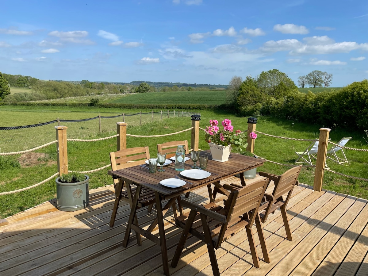 An outdoor dining area is set on a wooden deck, featuring a table and six folding chairs. A flower pot holds pink blooms, and the green countryside is visible in the background, showcasing distant trees and fields under a clear blue sky.