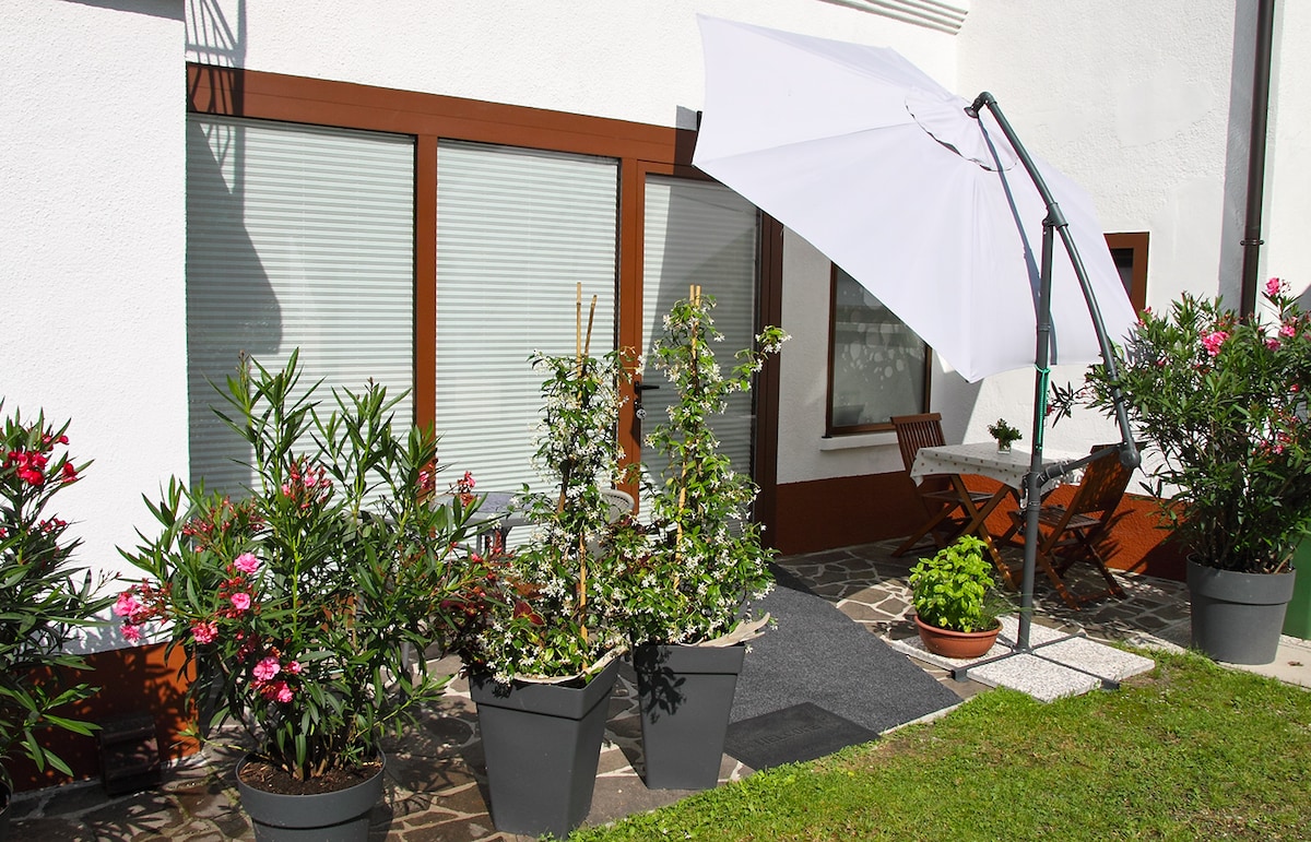 A welcoming outdoor area is featured, showcasing a patio with a small table and two chairs. Potted plants and shrubs enhance the space, complemented by a large umbrella providing shade. The entrance is framed by large windows and decorative flowers, creating a serene environment.
