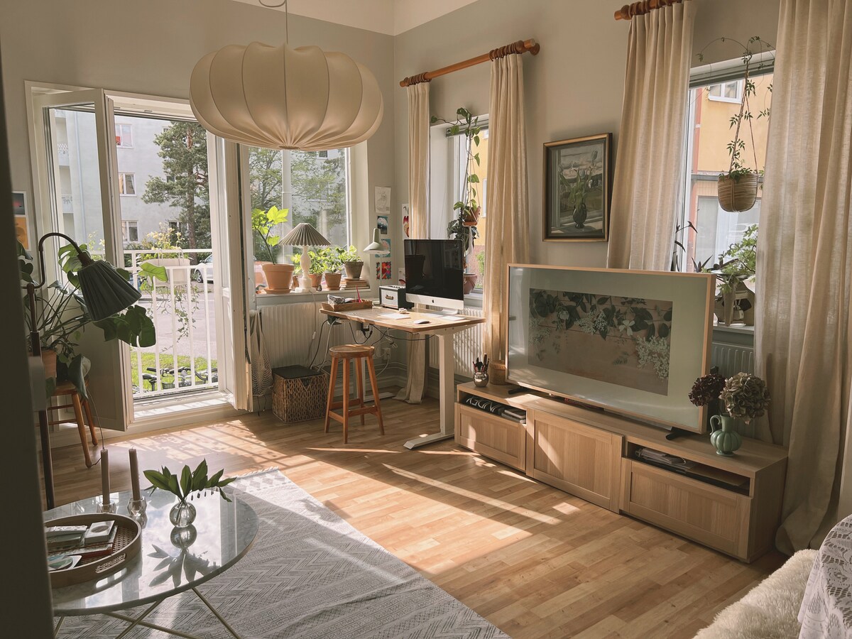 A bright living room is highlighted by large windows that offer natural light and views of greenery outside. A computer workstation is positioned beside a low wooden cabinet. Indoor plants are placed on various surfaces, and a round glass coffee table adds to the inviting decor.