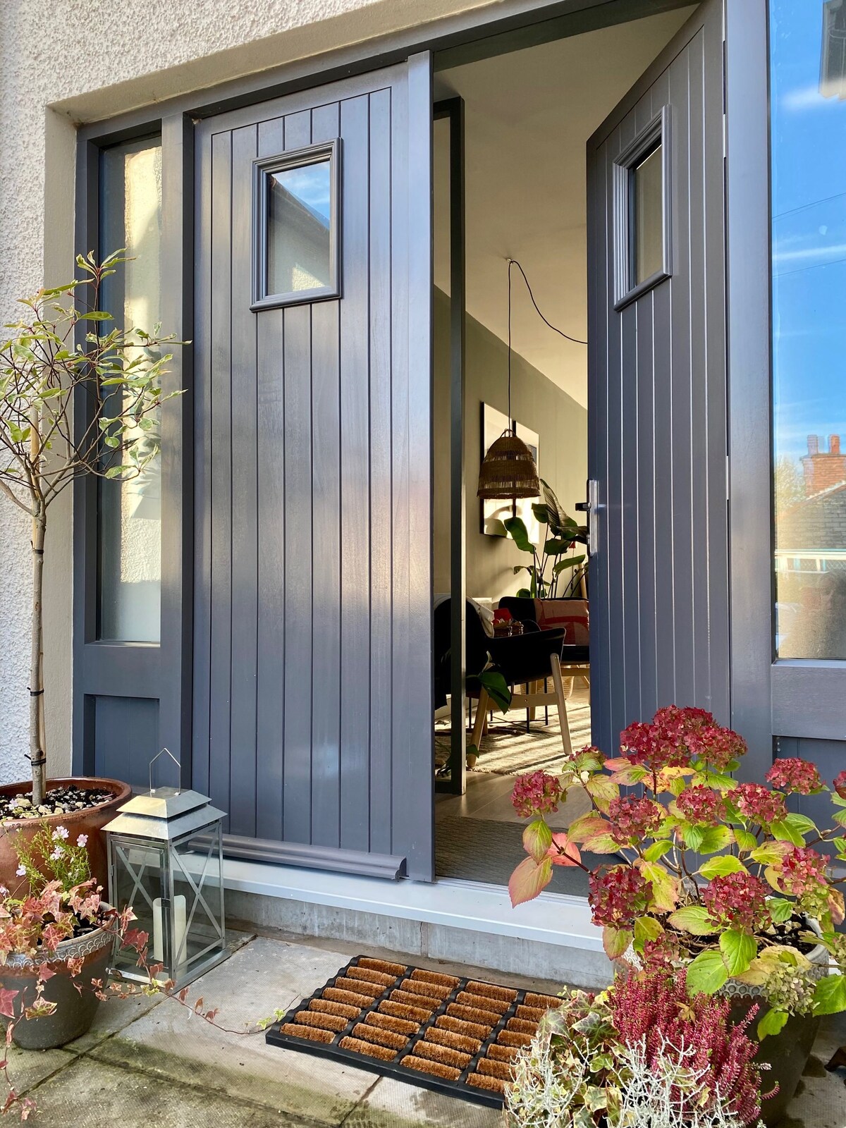 A welcoming entrance is framed by a grey double door, featuring two small windows. Potted plants and a lantern adorn the entryway, while a doormat suggests a neat and inviting space beyond. The interior is partially visible, hinting at a cozy atmosphere.