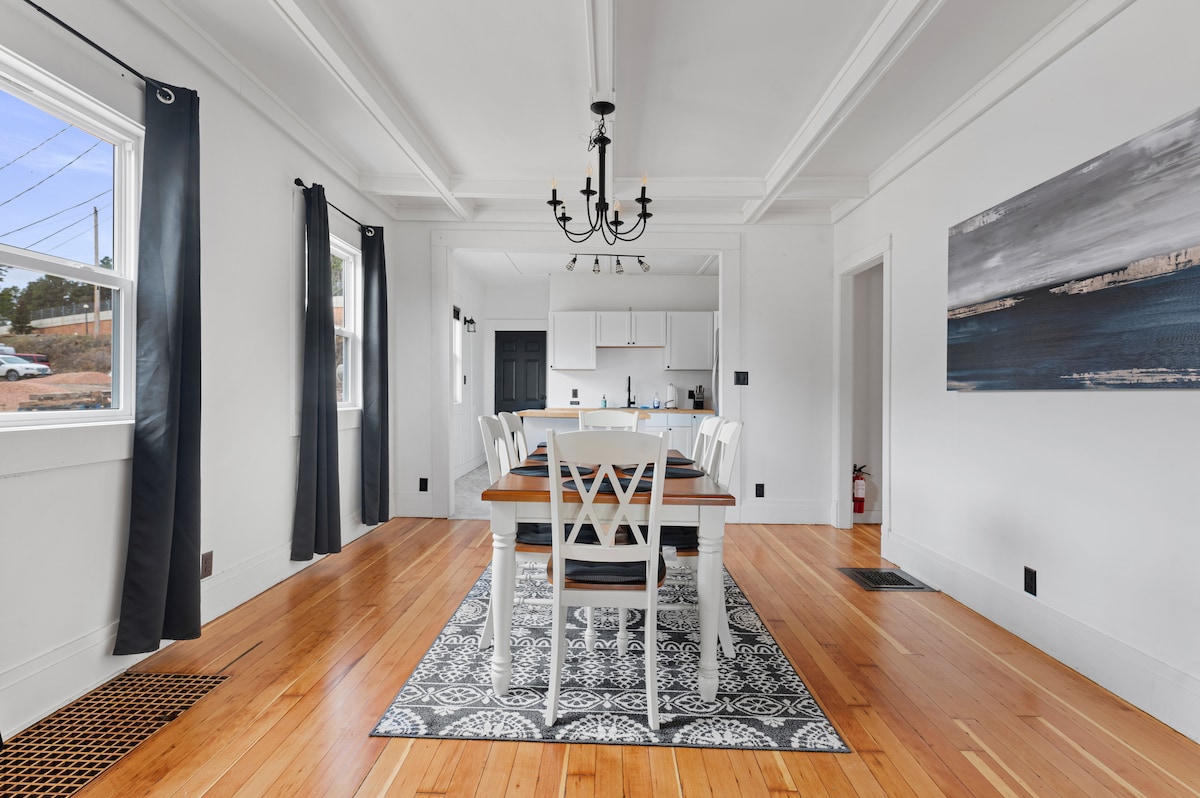 A bright dining area features a large wooden table with seating for six, complemented by two additional chairs at the counter. The room showcases hardwood floors, white walls, and a central chandelier, while natural light enters through two windows adorned with dark curtains.