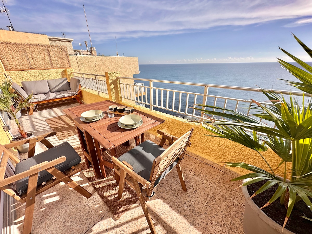 A large terrace is featured with a wooden dining table set for two, complemented by a cozy seating area. Potted plants frame the space, and unobstructed views of the Mediterranean Sea stretch beyond the railing, showcasing clear blue skies.