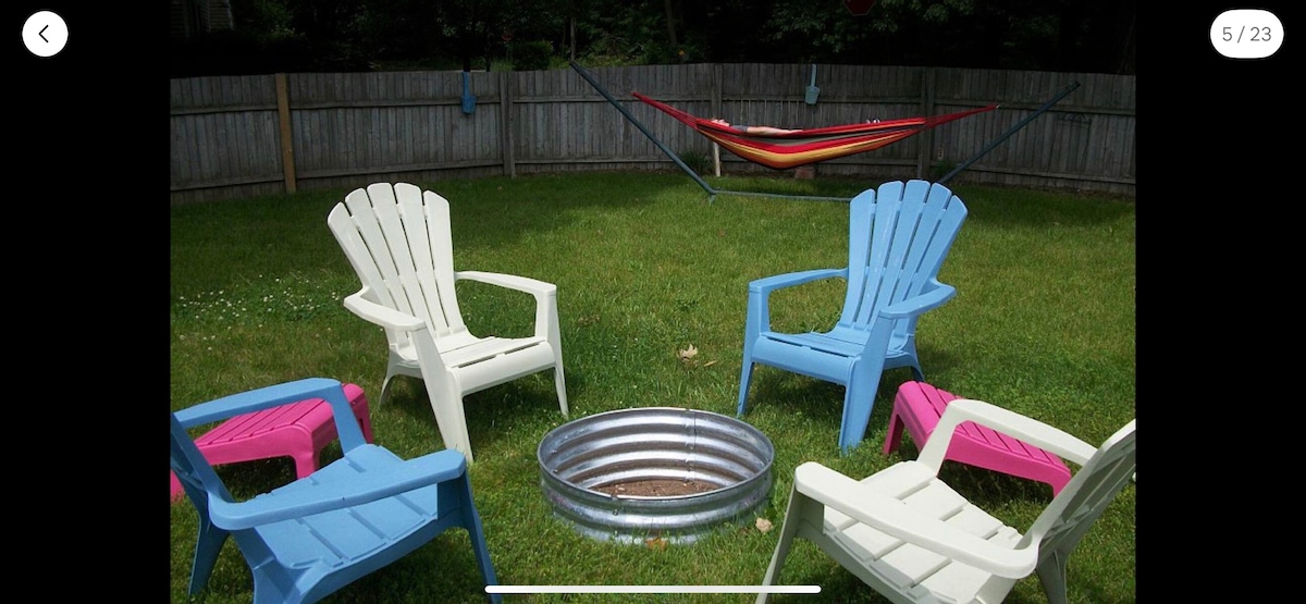 A fenced backyard is featured, showcasing a fire pit surrounded by four adirondack chairs in soft colors. A hammock is suspended between two supports in the background, adding a relaxed vibe to the outdoor space.