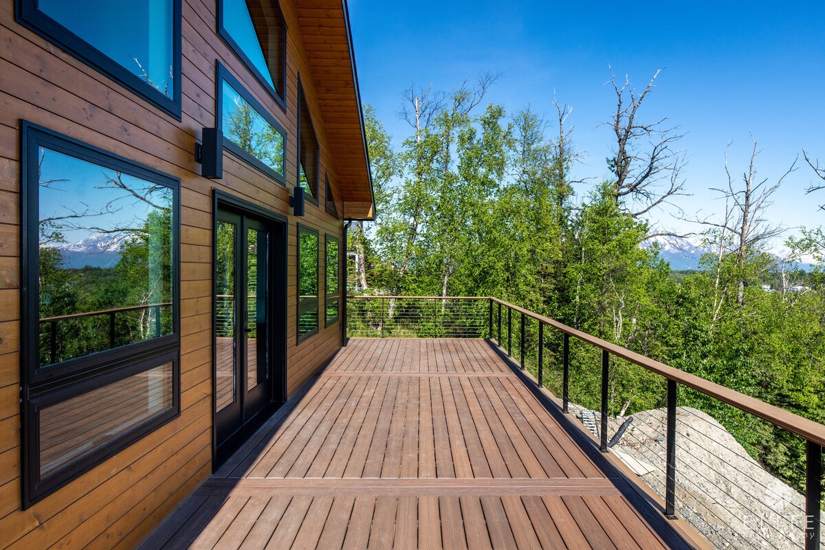 A spacious outdoor deck features wooden flooring and glass railings, providing unobstructed views of the surrounding trees and distant mountains. Large windows on the adjacent wall reflect natural light, creating a bright and open atmosphere. The deck offers ample room for relaxation or gatherings.