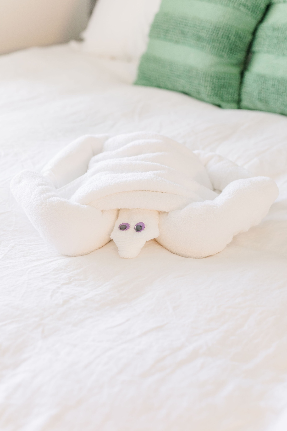 A creatively folded towel resembles a turtle, featuring soft, white fabric and two purple googly eyes. This charming towel art is placed on a neutral-colored bedspread, adding a playful touch to the sleeping area.