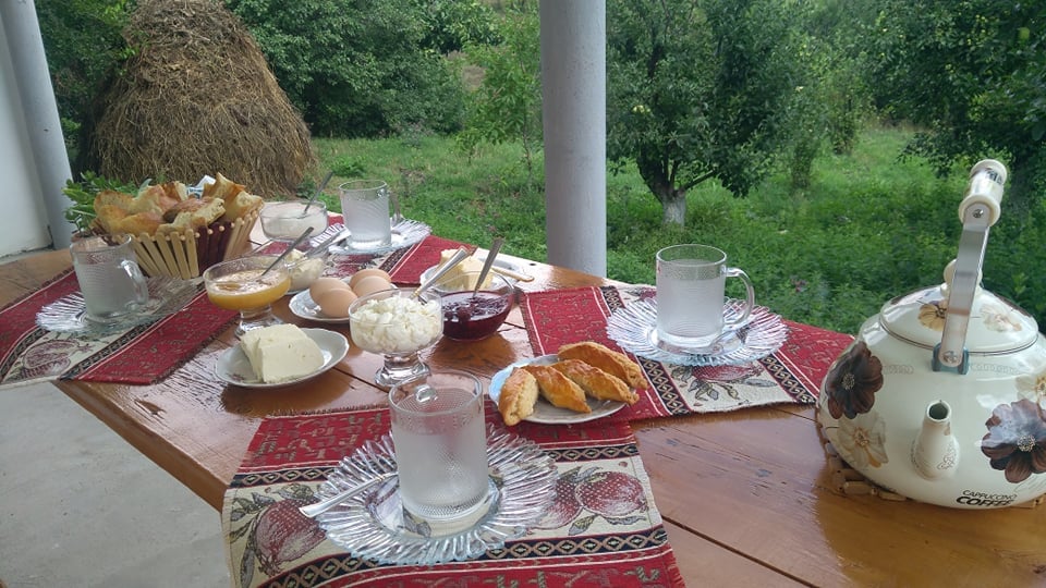 A table is set outdoors, featuring a variety of breakfast items, including pastries, eggs, and dairy products. Glasses of water are placed alongside dishes of jam and honey. The scene is complemented by a lush green backdrop, creating a peaceful dining atmosphere.