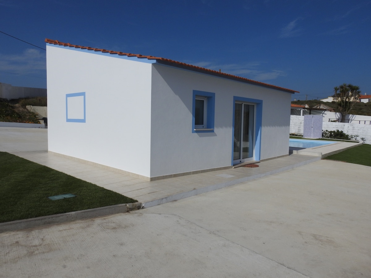A modern house with a clean white exterior and blue trim is pictured, featuring large glass doors that open towards an outdoor area. The surrounding space includes well-maintained grass and a glimpse of a swimming pool in the background.