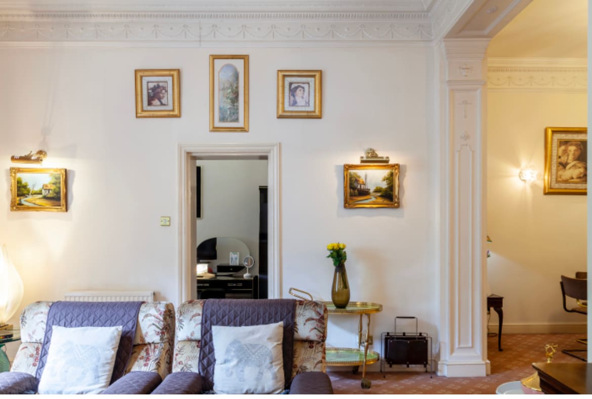 A stylish living area features a comfortable sofa adorned with decorative pillows. Framed artwork and photographs hang on the light-colored walls, complemented by elegant sconces. An open doorway leads to another room, creating an inviting flow in the space.