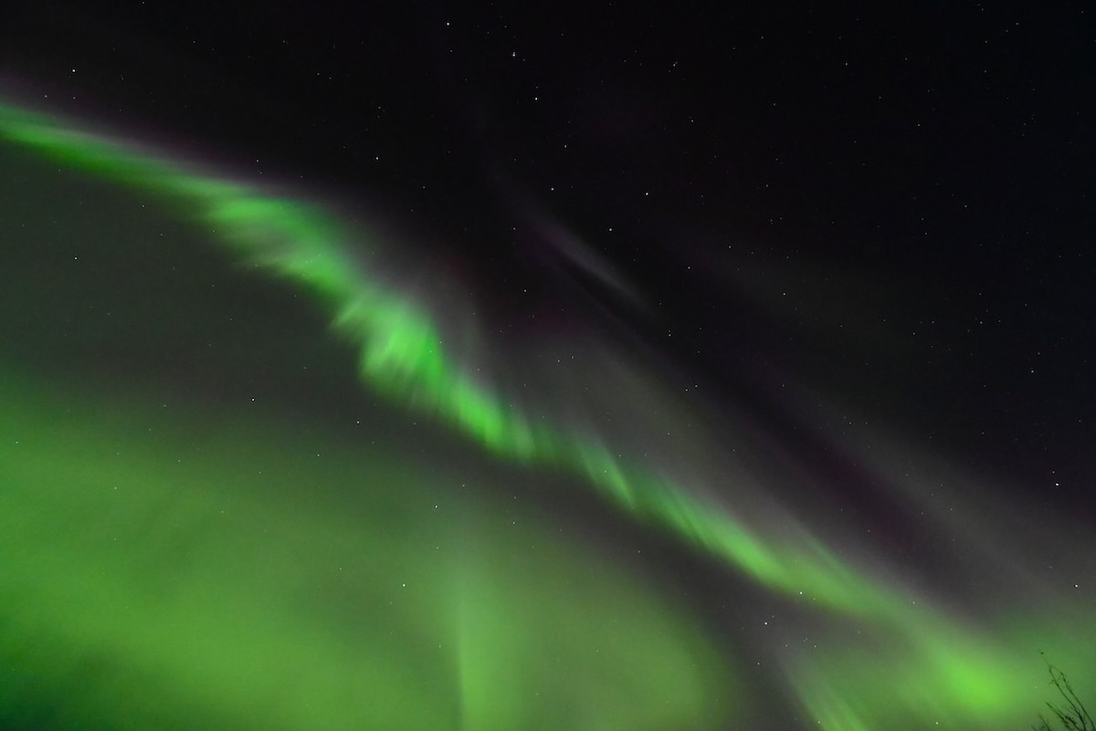 A display of the Northern Lights illuminates the night sky with vibrant green hues. Wispy patterns sweep across the dark backdrop, creating a mesmerizing spectacle of natural light. Stars are faintly visible, adding depth to the enchanting nighttime scene.