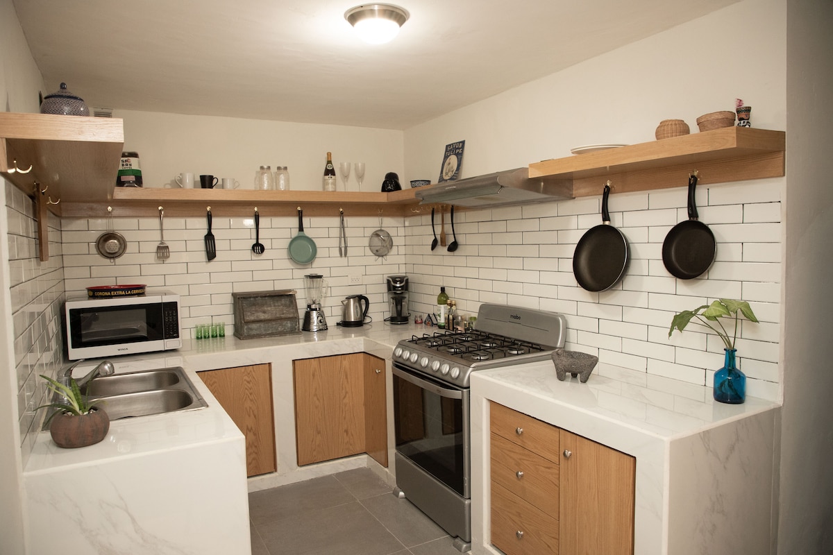 The kitchen features wooden shelves displaying various kitchen items. A gas stove and a microwave are positioned on the countertop, alongside a stainless steel sink. Pans hang from a rack above, while a small plant adds a touch of greenery to the space.