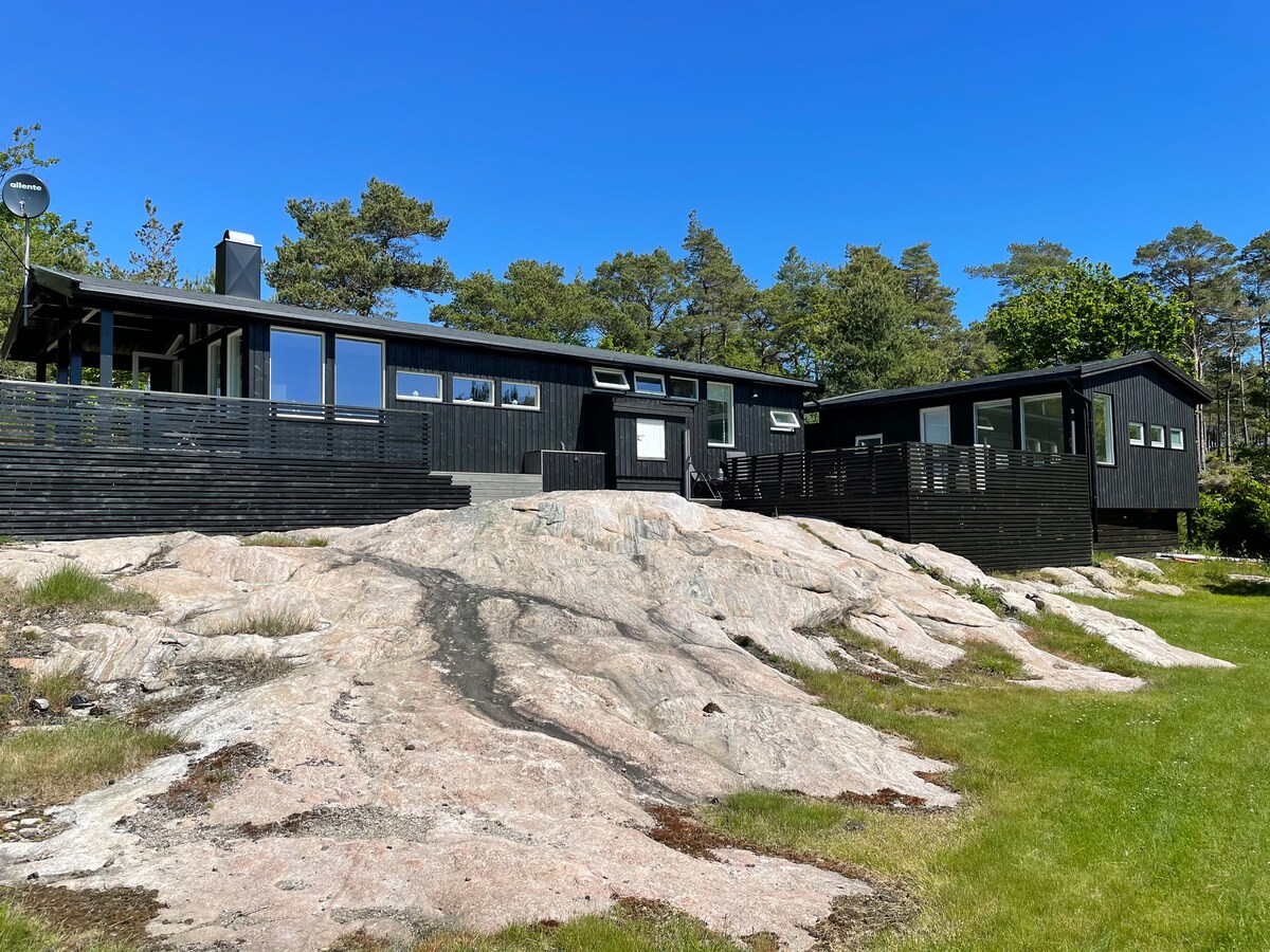 A modern black cabin is positioned on a rocky landscape, framed by lush greenery and a clear blue sky. The structure features large windows and a spacious deck, inviting outdoor relaxation while blending with the natural surroundings.