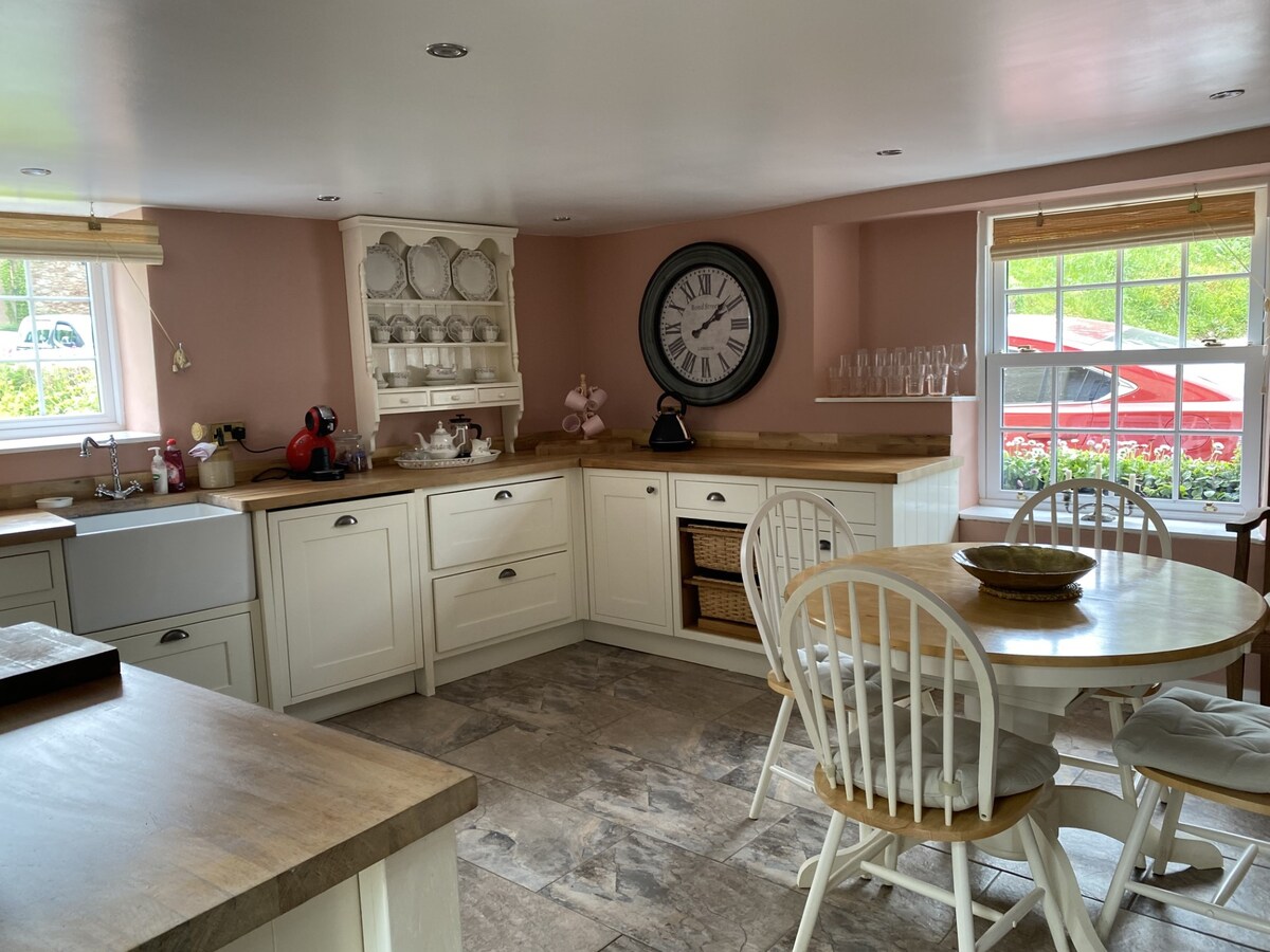 A well-equipped kitchen features light cabinetry and a rounded dining table with four chairs. A large clock is mounted on a pink wall, and natural light filters in through a window, highlighting the sink area and cooking appliances.