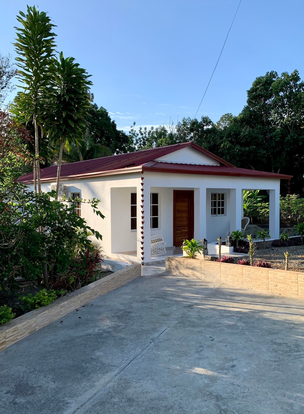 A charming one-story cabaña is surrounded by lush greenery, featuring a red roof and white exterior. A covered porch offers a welcoming entrance, complemented by a pathway leading through a well-maintained garden setting. Large windows provide natural light and enhance the inviting appearance.