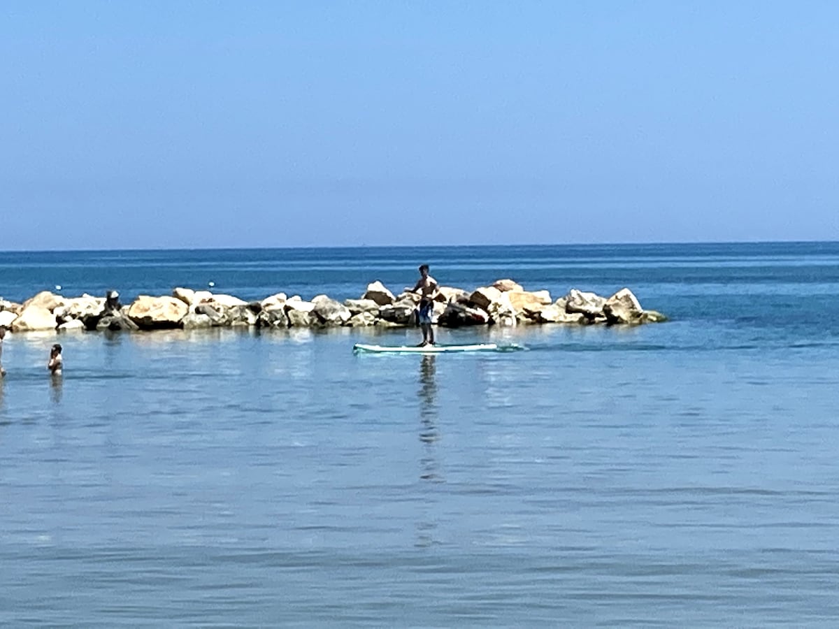 A serene ocean scene captures a person paddleboarding near a rocky shore. Gentle waves ripple across the calm water, reflecting the clear blue sky. In the distance, other individuals can be seen enjoying a leisurely swim, enhancing the tranquil coastal atmosphere.