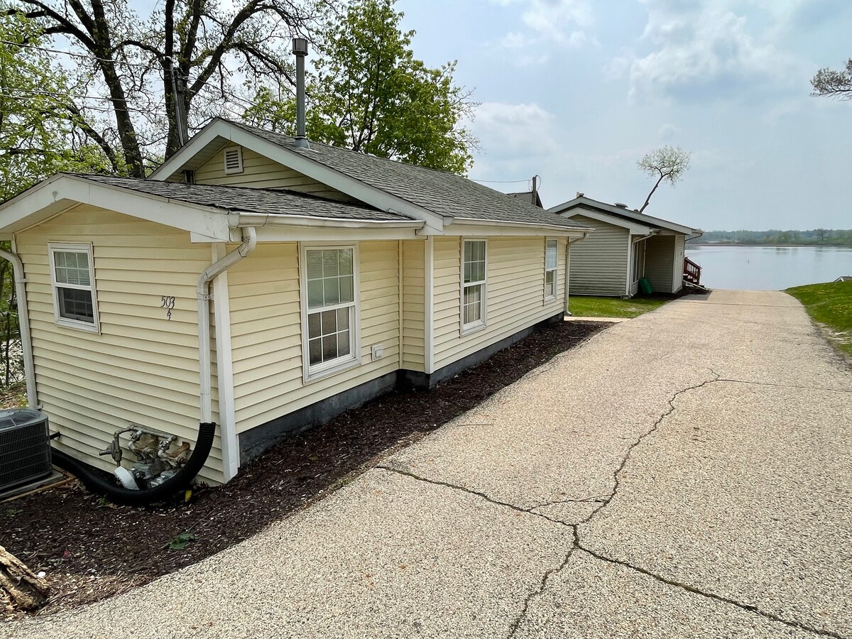 The exterior of a small house is shown, featuring light yellow siding and multiple windows. A gravel drive leads to the home, with a serene view of the water nearby. Trees and greenery are visible, contributing to the peaceful setting.
