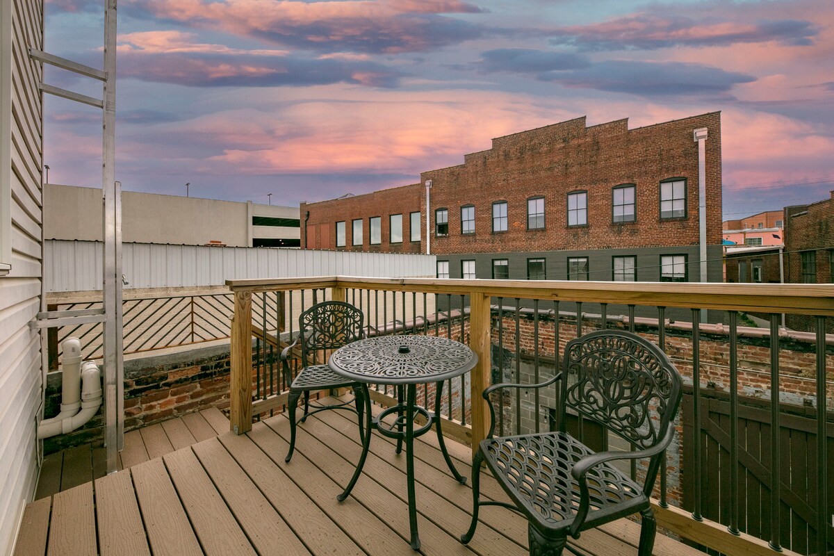 A rear deck features a small table and two ornate chairs, providing a cozy outdoor space. The deck is surrounded by brick buildings, with a colorful sky visible above, suggesting a tranquil setting for relaxation.