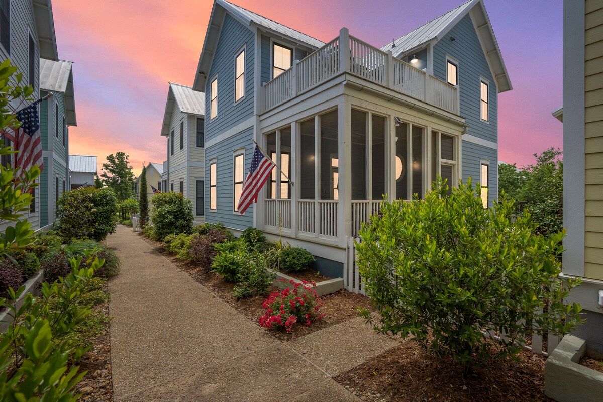A welcoming exterior of a two-story blue house is showcased with a screened-in porch and an American flag. Lush greenery and flower beds line a pebble path leading to the entrance, creating a calm and inviting environment at twilight.